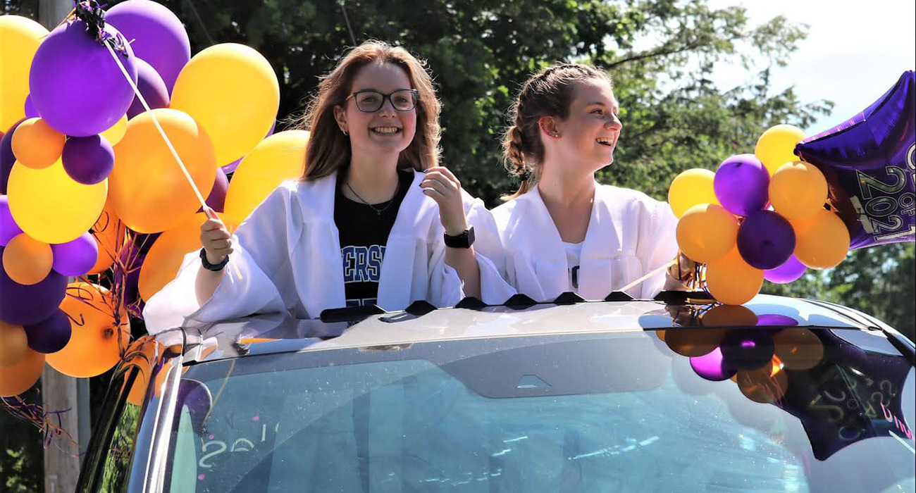 two high school students riding in a car wearing their robes and holding balloons